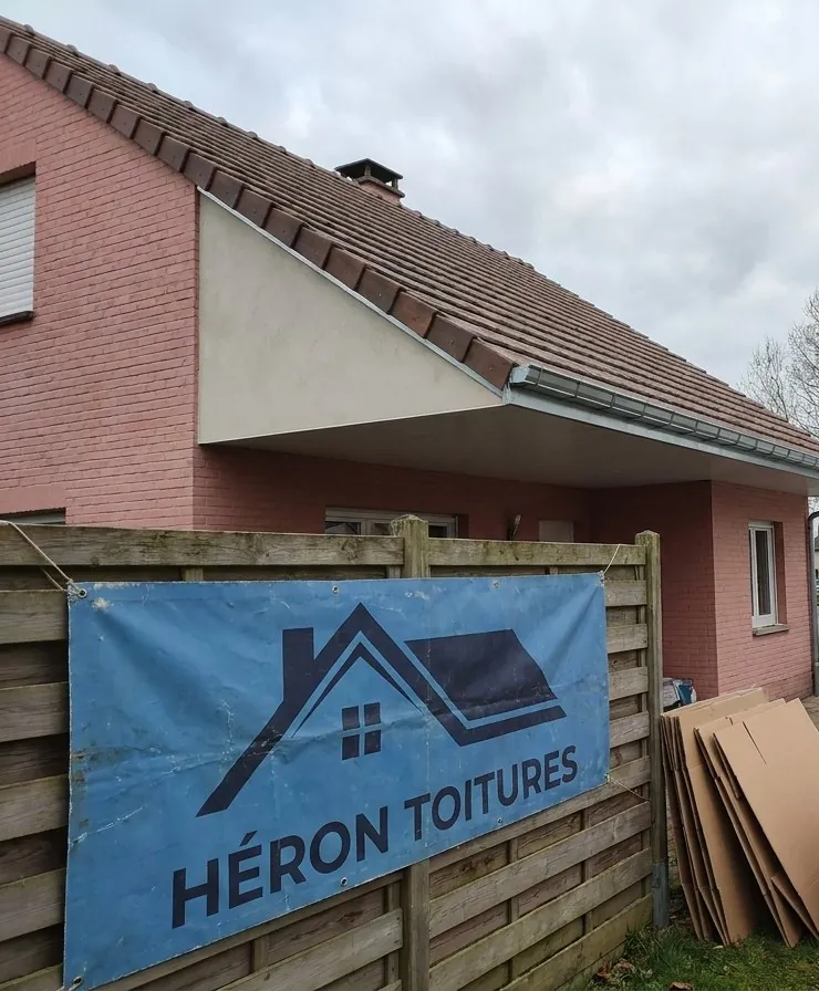 Bâche bleue Héron Toitures accrochée à la clôture d'un pavillon en brique rose pendant des travaux de couverture