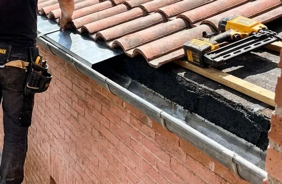 Main d'un couvreur zingueur posant une bande de zinc sous le premier rang de tuiles canal, gouttière neuve en dessous, Saint-Jean